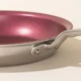 A close-up of a metallic frying pan with a red-hued interior and a riveted handle.