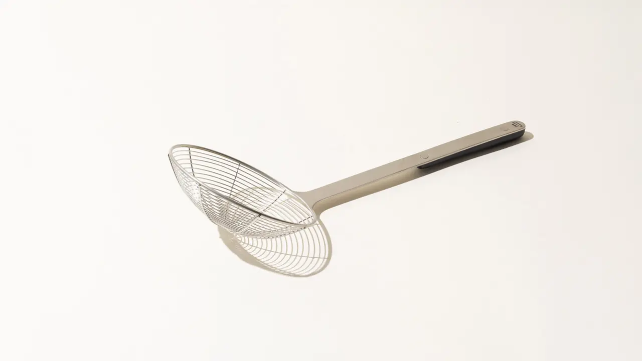 A stainless steel kitchen strainer with a long handle is displayed against a plain background.