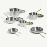 A variety of stainless steel cookware pieces with gold handles are arranged on a light background.
