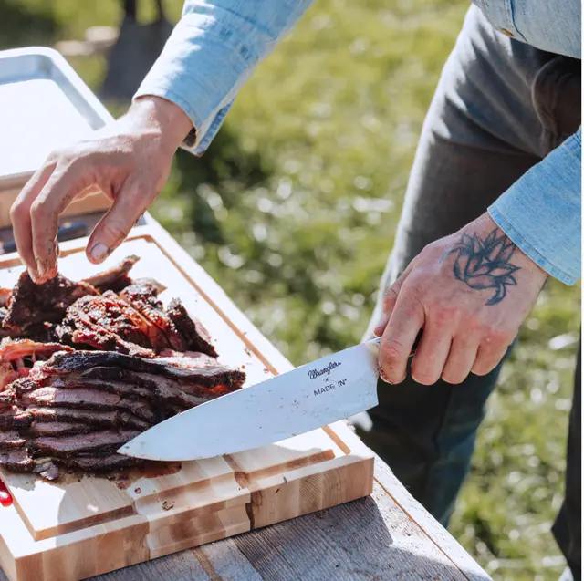 A person is slicing grilled meat on a wooden cutting board while using a large knife outdoors.
