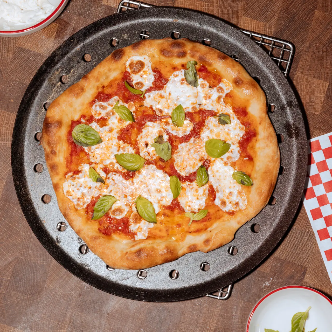 A freshly baked pizza with melted cheese and basil leaves is presented on a round black baking tray on a wooden surface.