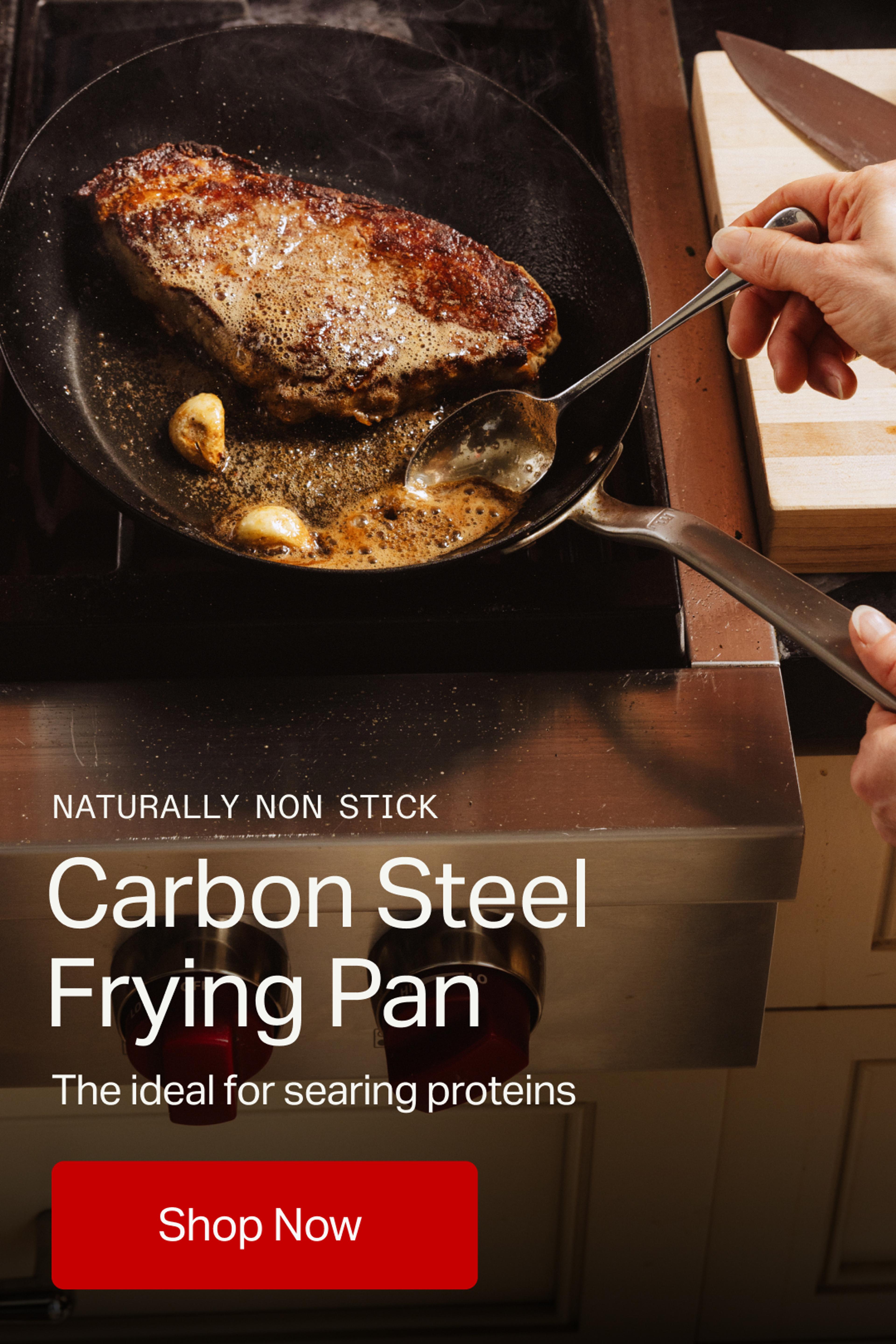 A hand holds a spatula while cooking a seared steak with garlic in a carbon steel frying pan on a stovetop.