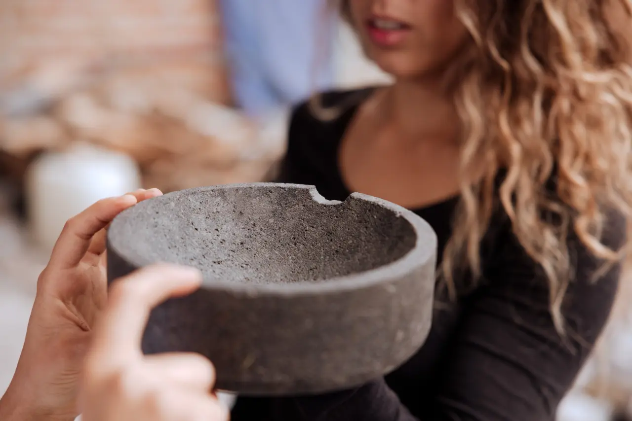 A person hands a grey, bowl-shaped concrete object to another person whose curly hair is partially in view.