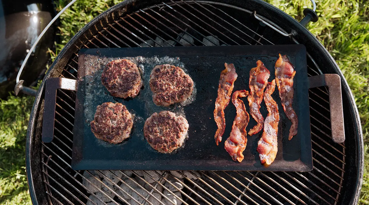 Hamburgers and bacon strips cook on a flat grill surface placed over a barbecue grill outdoors.