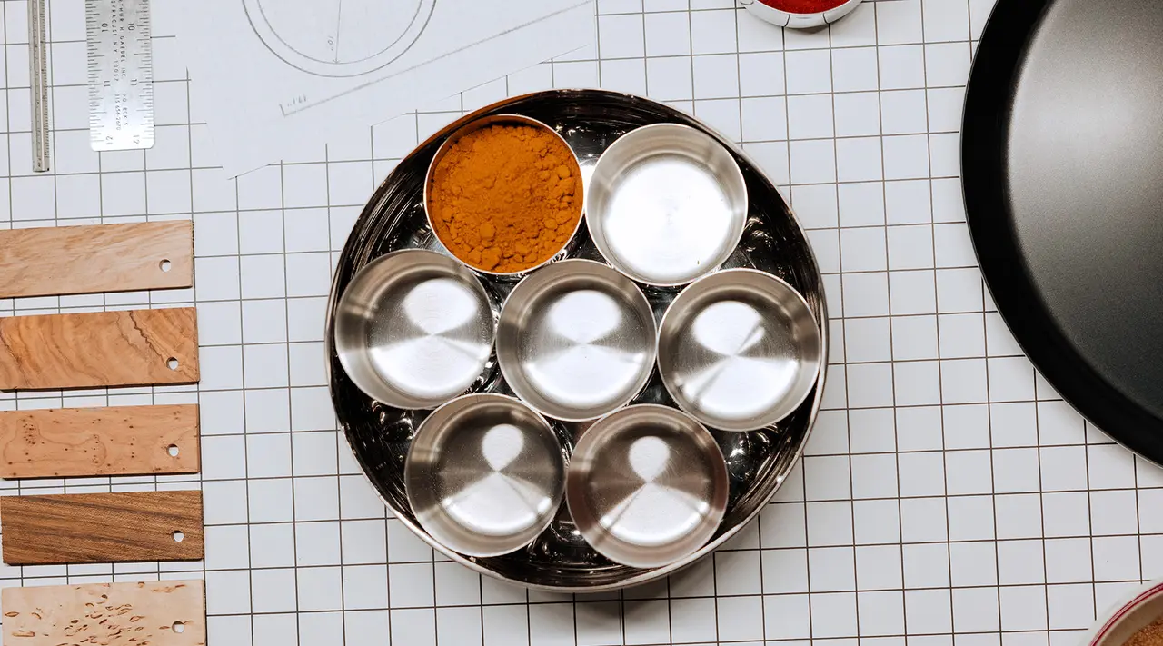 A spice palette with one compartment filled with turmeric lies on a patterned surface beside cooking utensils and a pan.