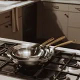 Stacked stainless steel frying pans with gold handles sit on a gas stove in a modern kitchen.