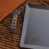 A rectangular, dark metal cooking pan with a textured handle that features the word "TEXAS" rests on a woven fabric surface.