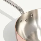 A shiny stainless steel saucepan with a copper exterior is shown with its handle.