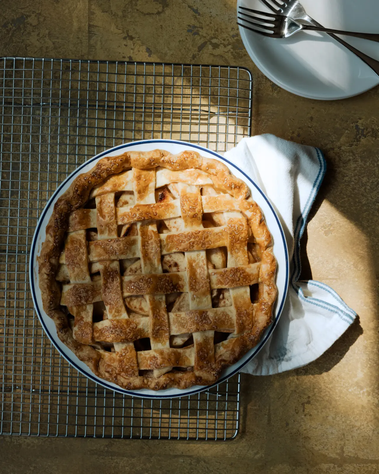 A freshly baked apple pie with a golden lattice crust sits on a cooling rack alongside a white napkin and silverware.