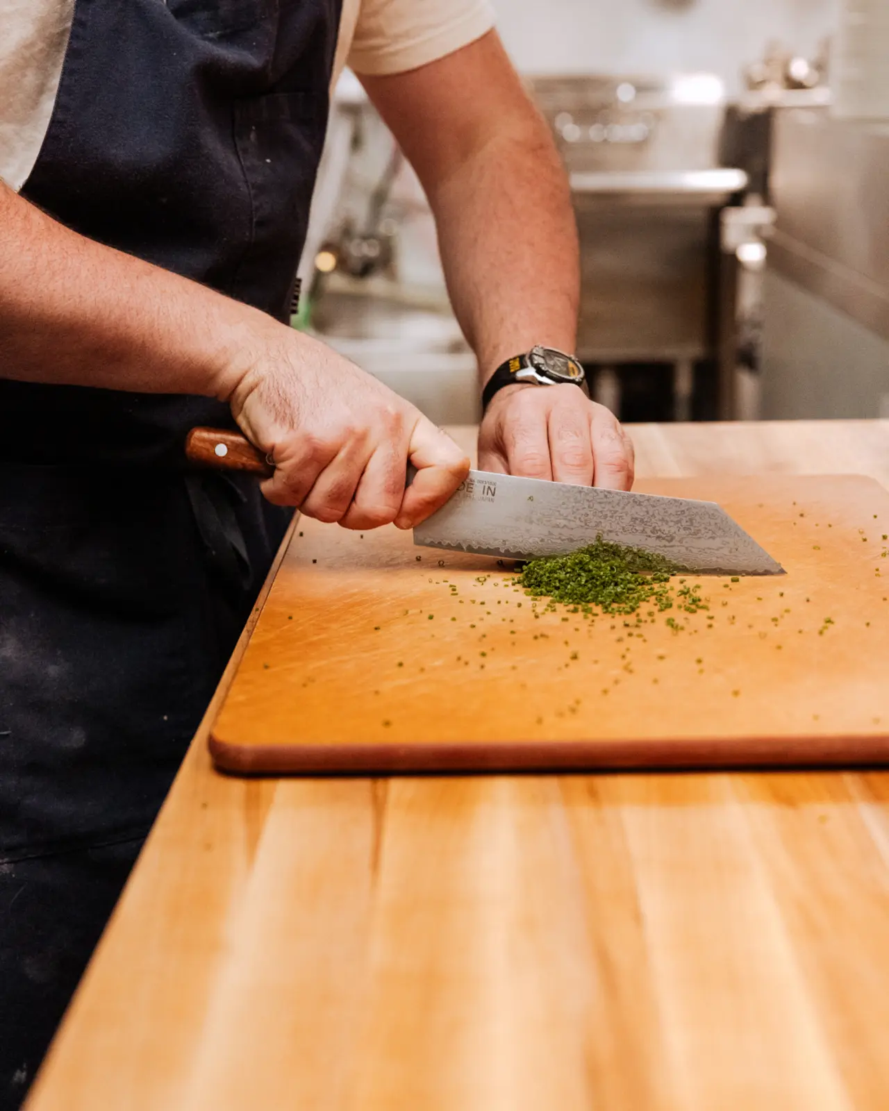 A person in an apron finely chops herbs on a wooden cutting board with a large chef's knife.