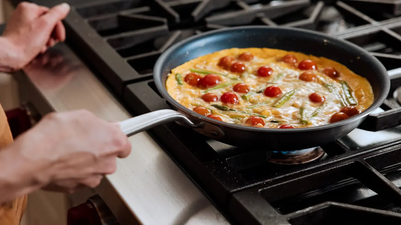 A person cooks an omelette with tomatoes and green beans in a skillet on a stove.