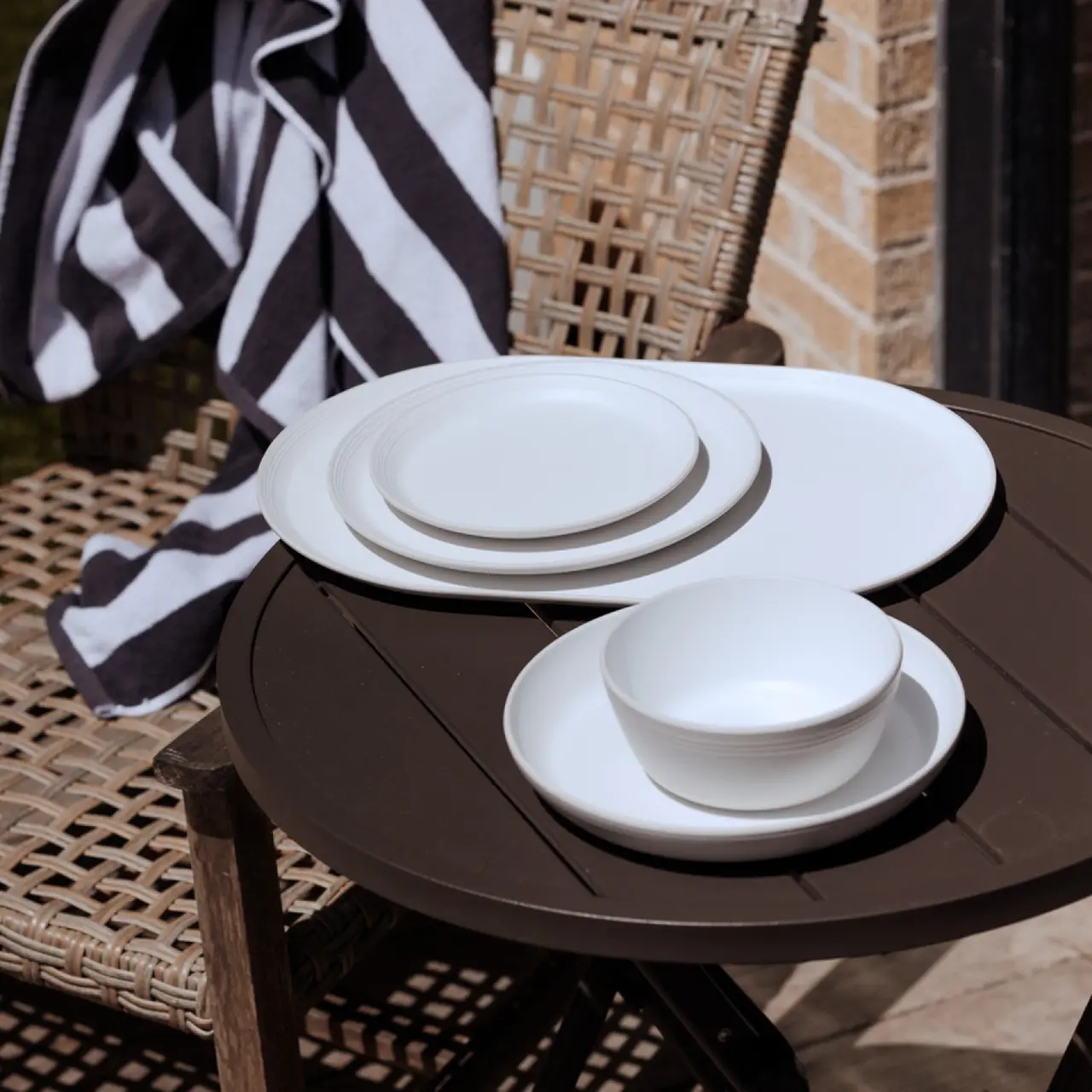 A set of white dishes is neatly stacked on a round outdoor table beside a draped striped towel and a wicker chair in the sunlight.