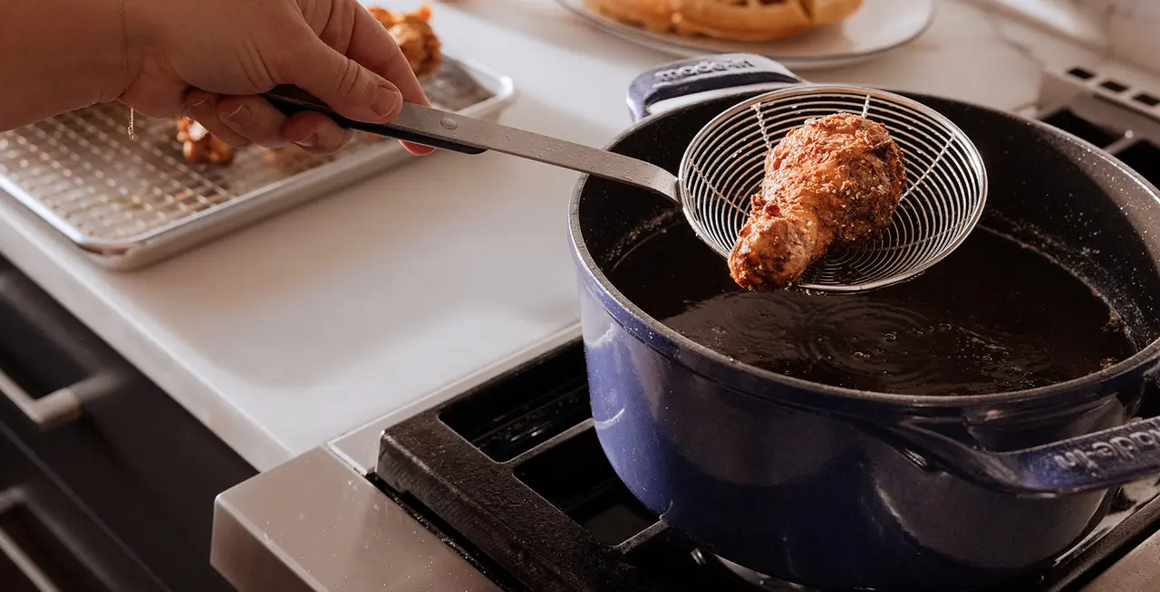 A person uses a metal utensil to lift a piece of fried chicken from a deep pot of oil onto a tray.