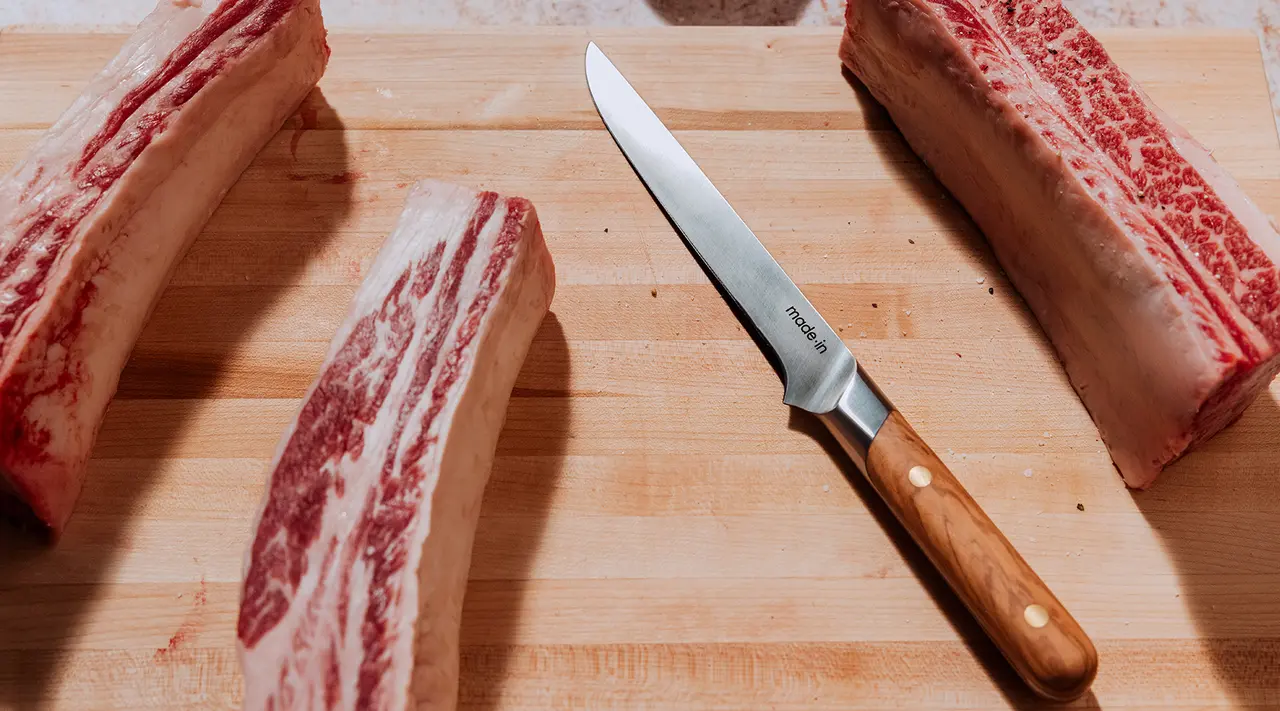 A sharp knife lies on a cutting board next to three thick cuts of raw meat with marbled fat.