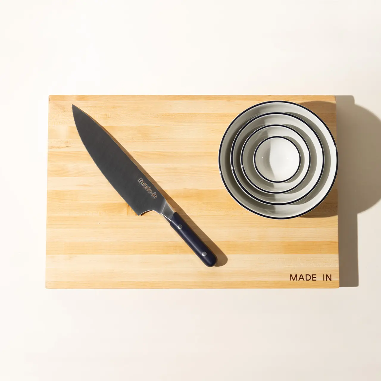 A chef's knife lies to the left of a stack of nested metal bowls on a wooden cutting board with the text "MADE IN" visible.