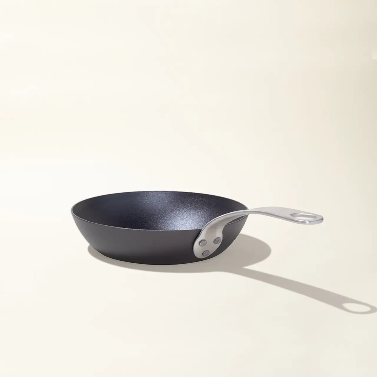 A black non-stick frying pan with a silver handle casts a shadow on a light surface.
