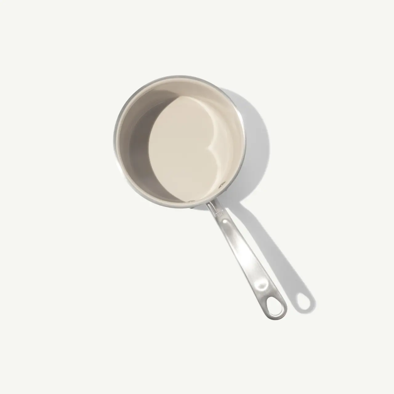 A simple stainless steel saucepan with a long handle sits against a light background, casting a subtle shadow.