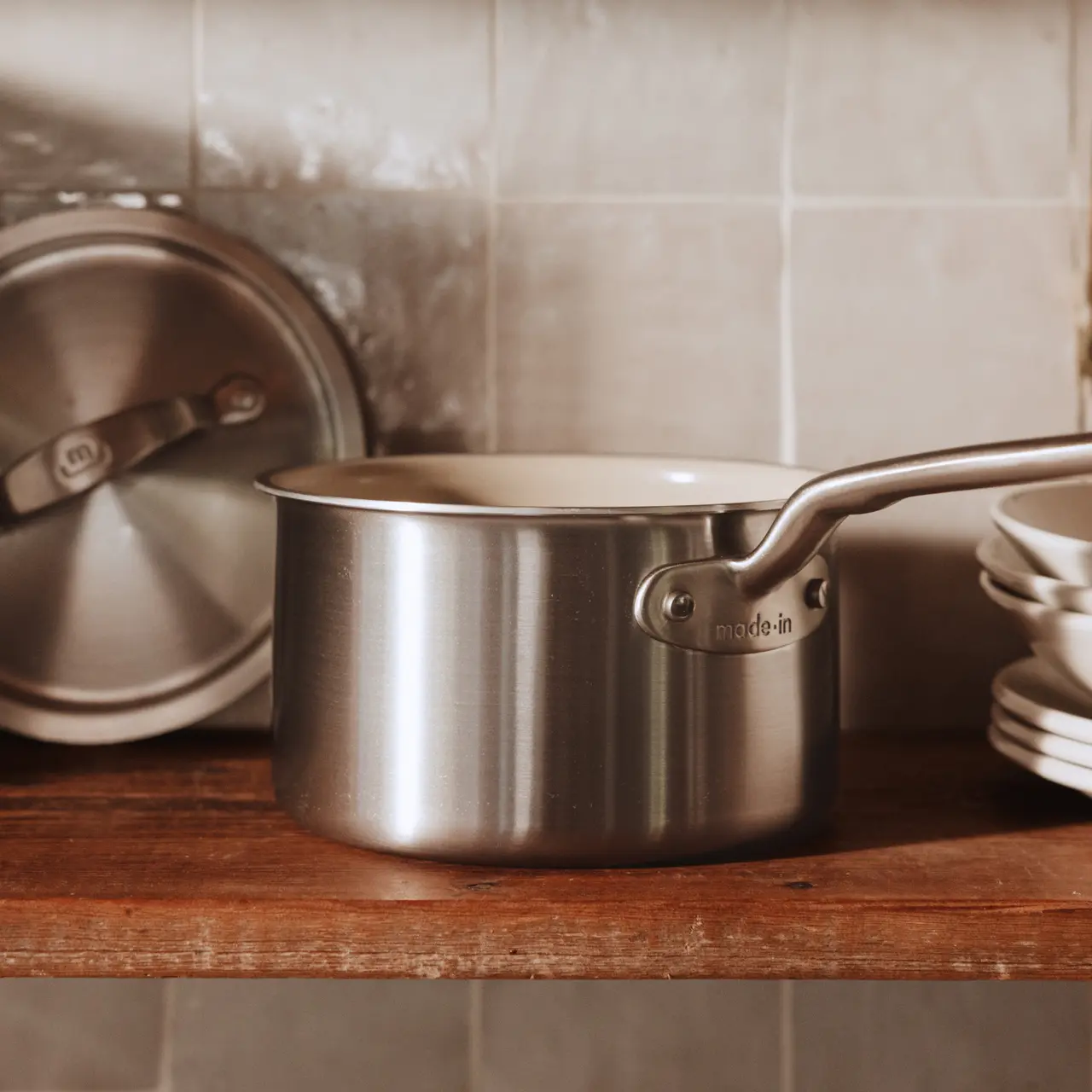 A sleek stainless steel saucepan with a lid rests on a wooden shelf, accompanied by additional cookware in the background.