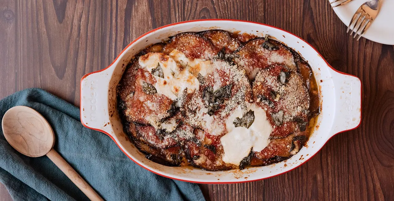 A baked dish featuring lasagna in a white casserole dish, placed on a wooden table with a wooden spoon and green cloth nearby.