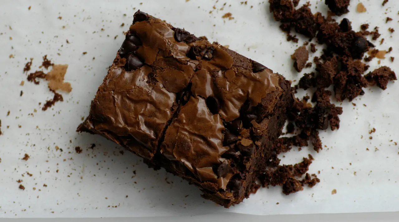 A close-up of a chocolate brownie with a cracked surface, surrounded by crumbs on a light background.