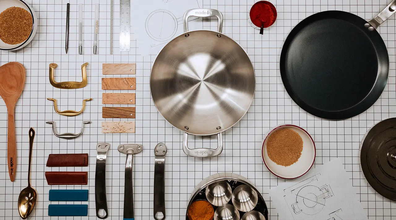 Various kitchen utensils and cookware are neatly arranged on a gridded surface, showcasing an assortment of items like pans, spoons, and measuring tools possibly for a design or culinary presentation.