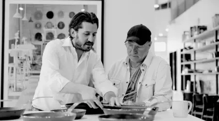 Two men, one younger with a beard and another older wearing a cap, attentively examine documents at a table with kitchenware around them.