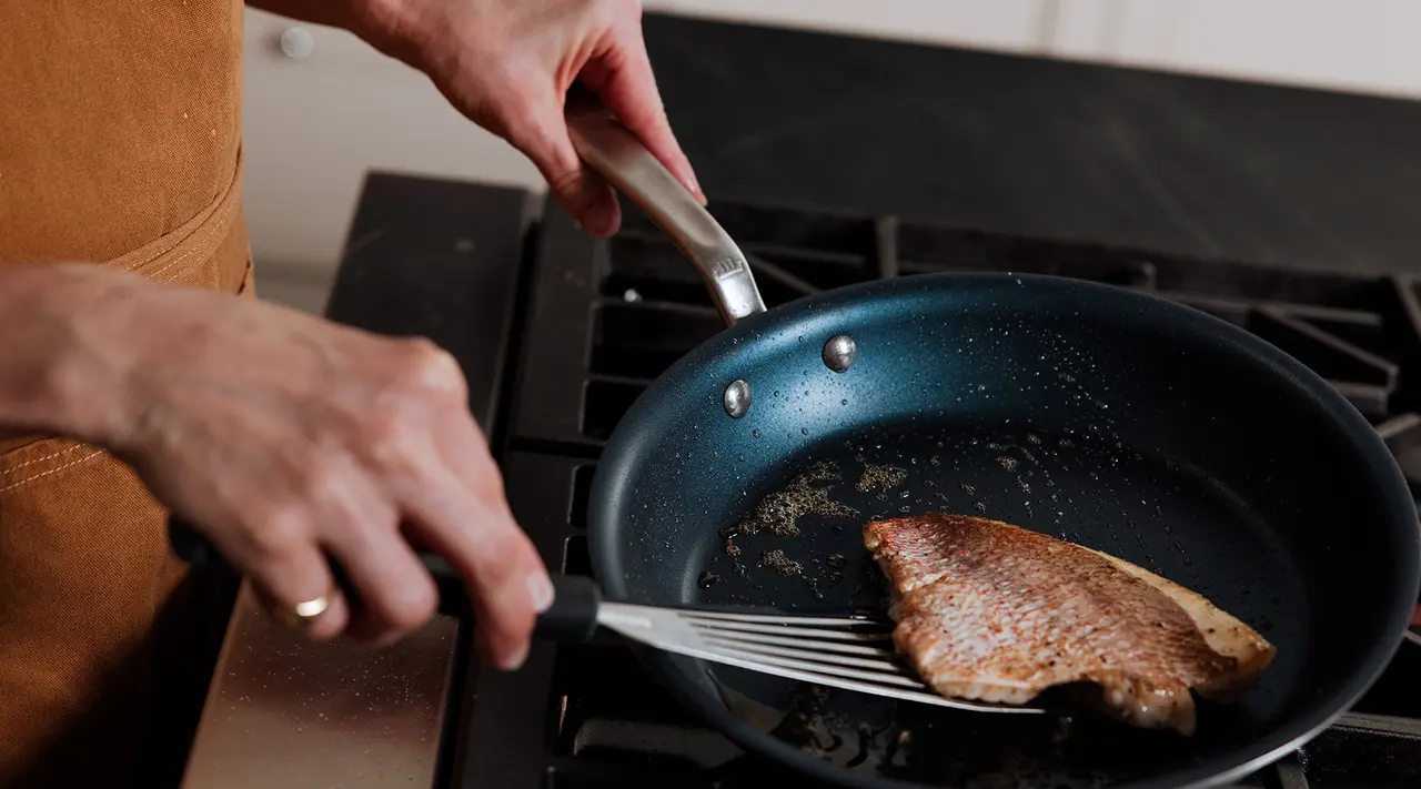 A person cooks a piece of fish in a skillet on a stove top.
