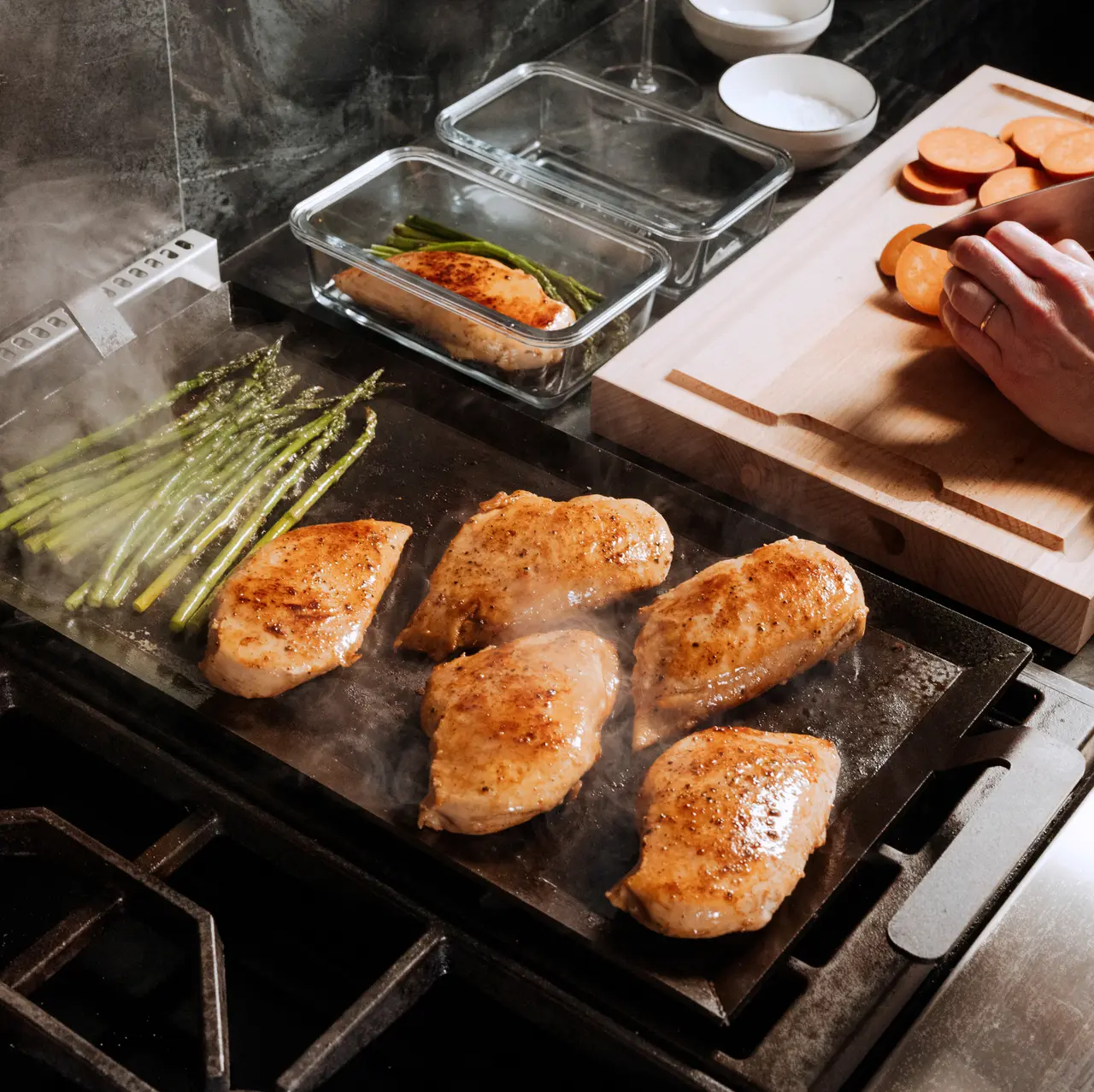 Chicken breasts are being seared on a stovetop griddle, surrounded by ingredients like asparagus, suggesting meal preparation.