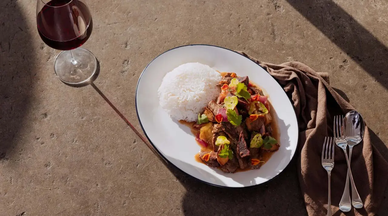 A plate of food with rice and a meaty stew paired with a glass of red wine and cutlery set on a table in sunlight.