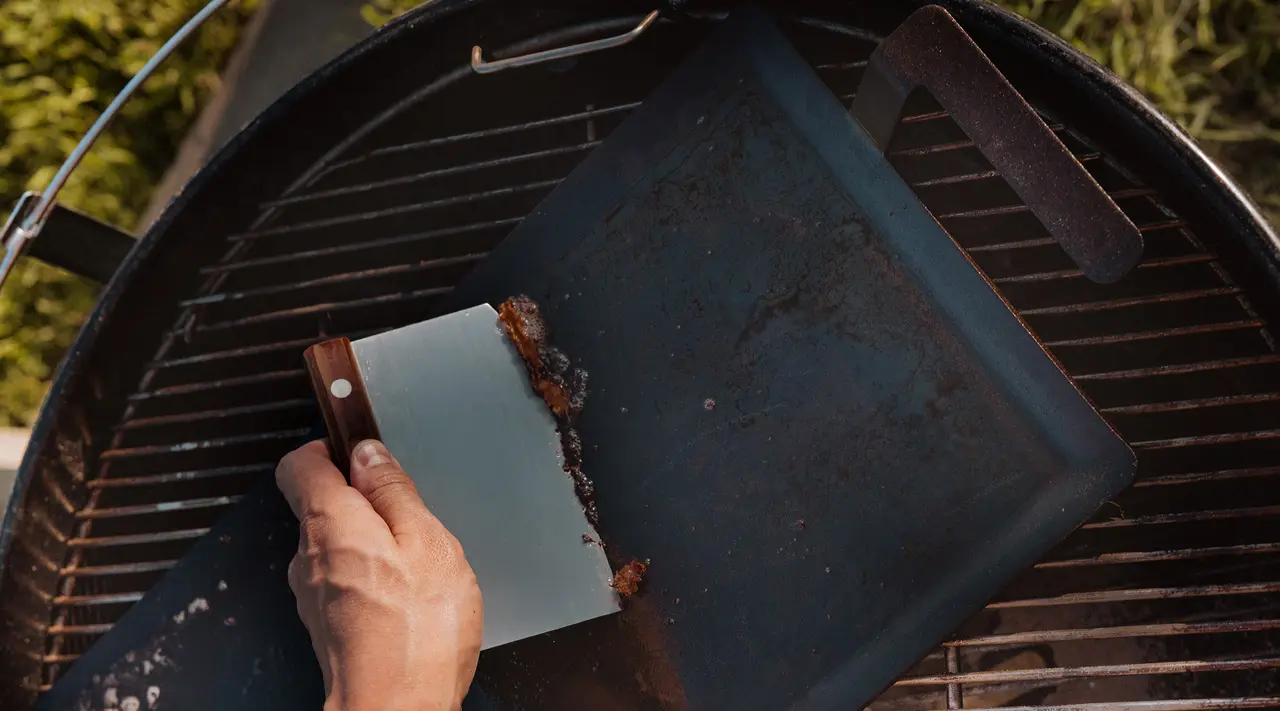 A person's hand holds a cleaver on a cutting board placed on a barbeque grill, with no visible food on the board.