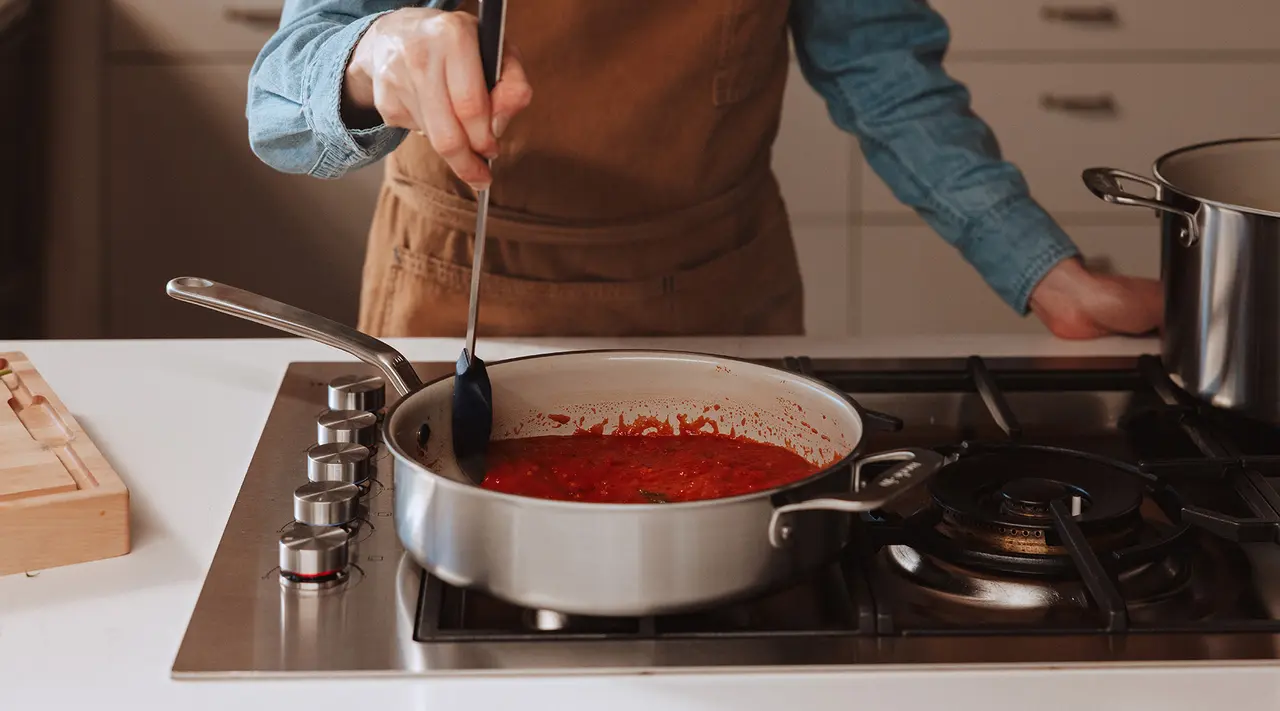 A person wearing an apron stirs a pot of red sauce on a gas stove.