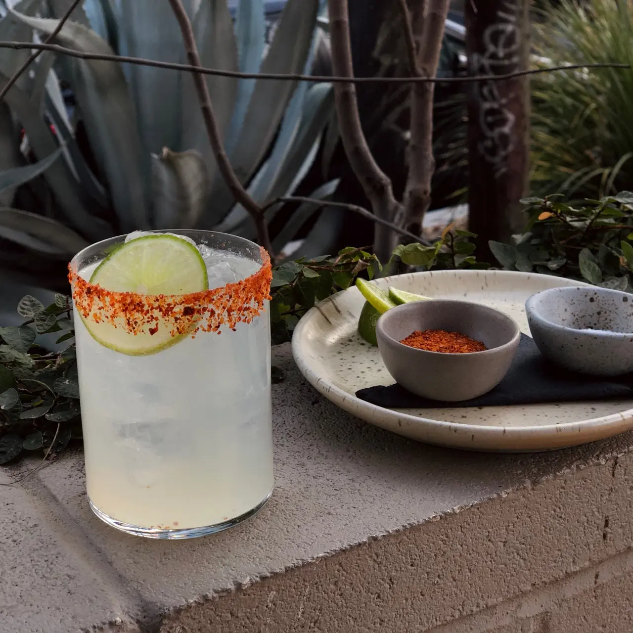 A refreshing drink with a rim coated in red seasoning sits beside a bowl of chili powder and lime wedges on an outdoor ledge with greenery in the background.