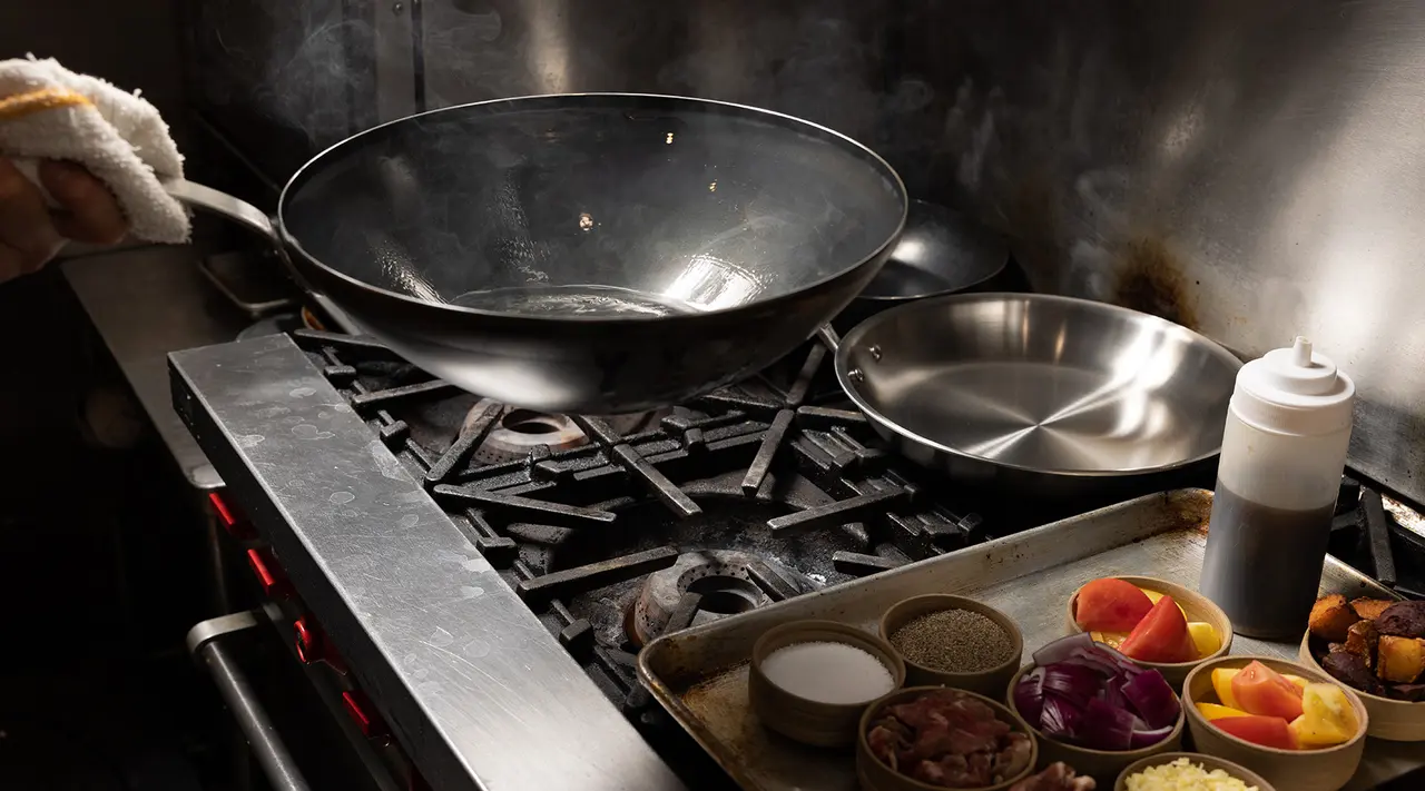 A chef's hand is visible holding a towel next to a smoking wok on a professional stove, surrounded by organized ingredients ready for cooking.