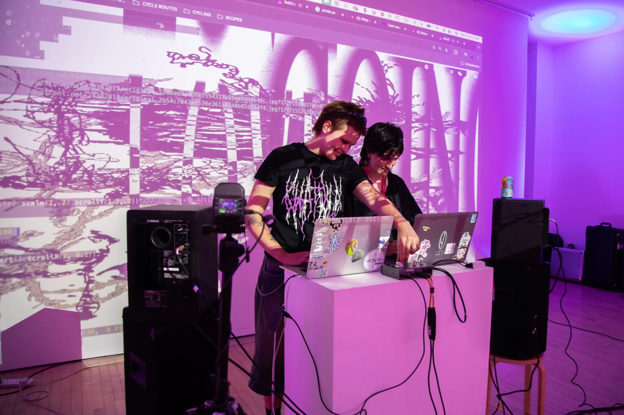 Two live coders stand at a desk with laptops while their visuals are projected behind them