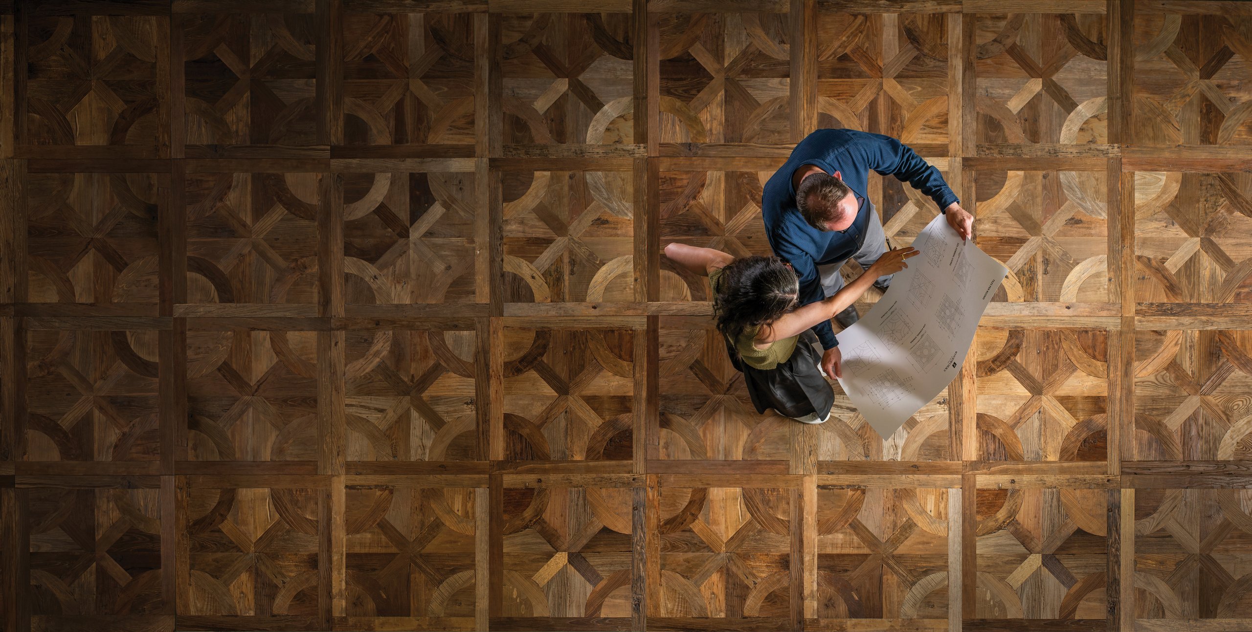 Two people reviewing drawings on a reclaimed parquet de Versailles floor, seen from above
