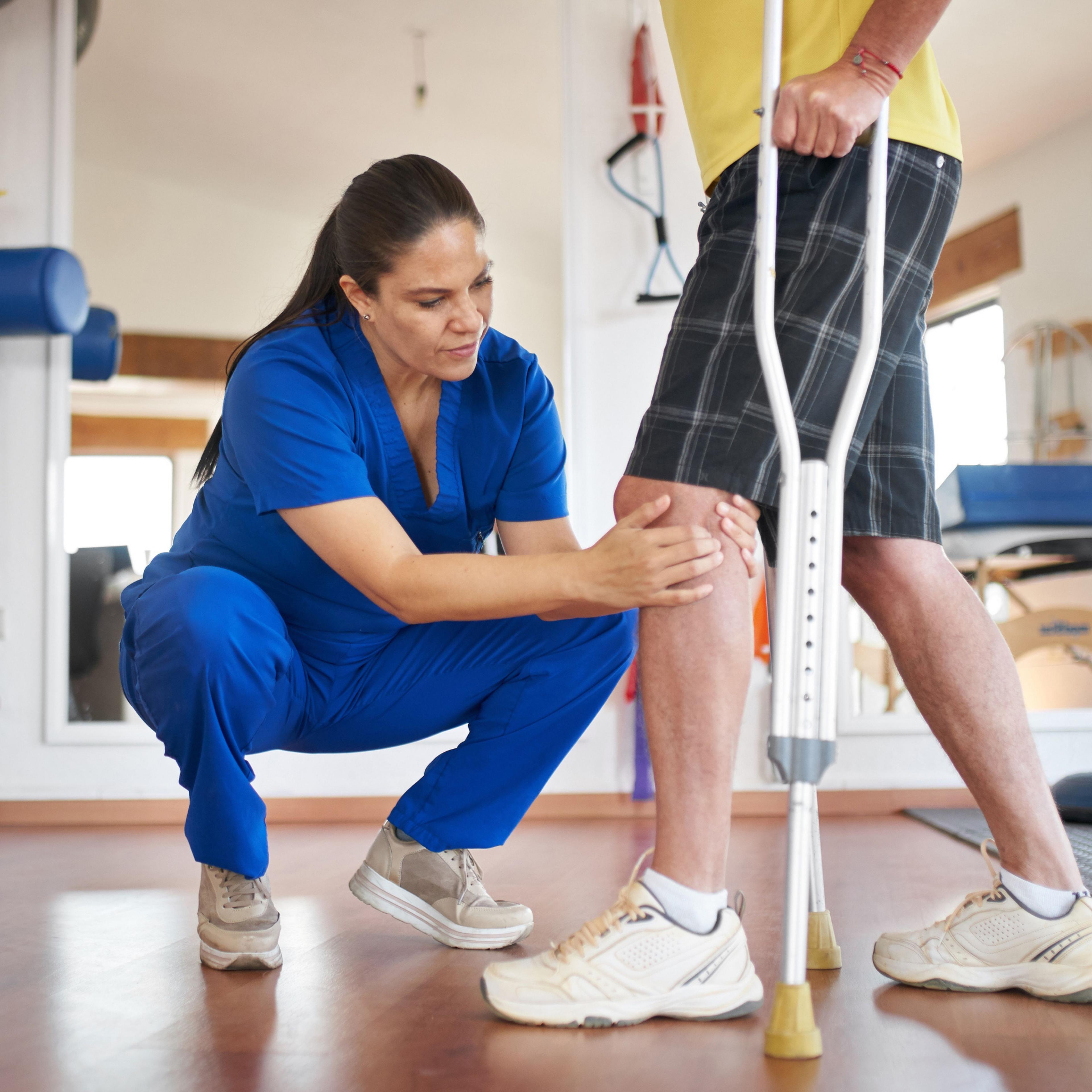 A HealthPro Heritage physical therapist examining a patient's knee while the patient uses a crutch