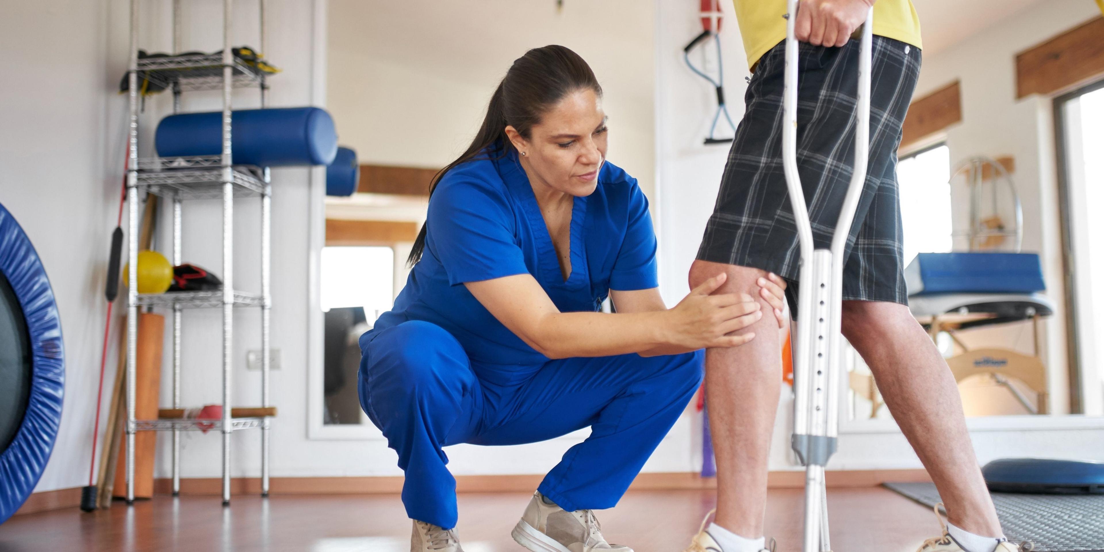 A HealthPro Heritage physical therapist examining a patient's knee while the patient uses a crutch