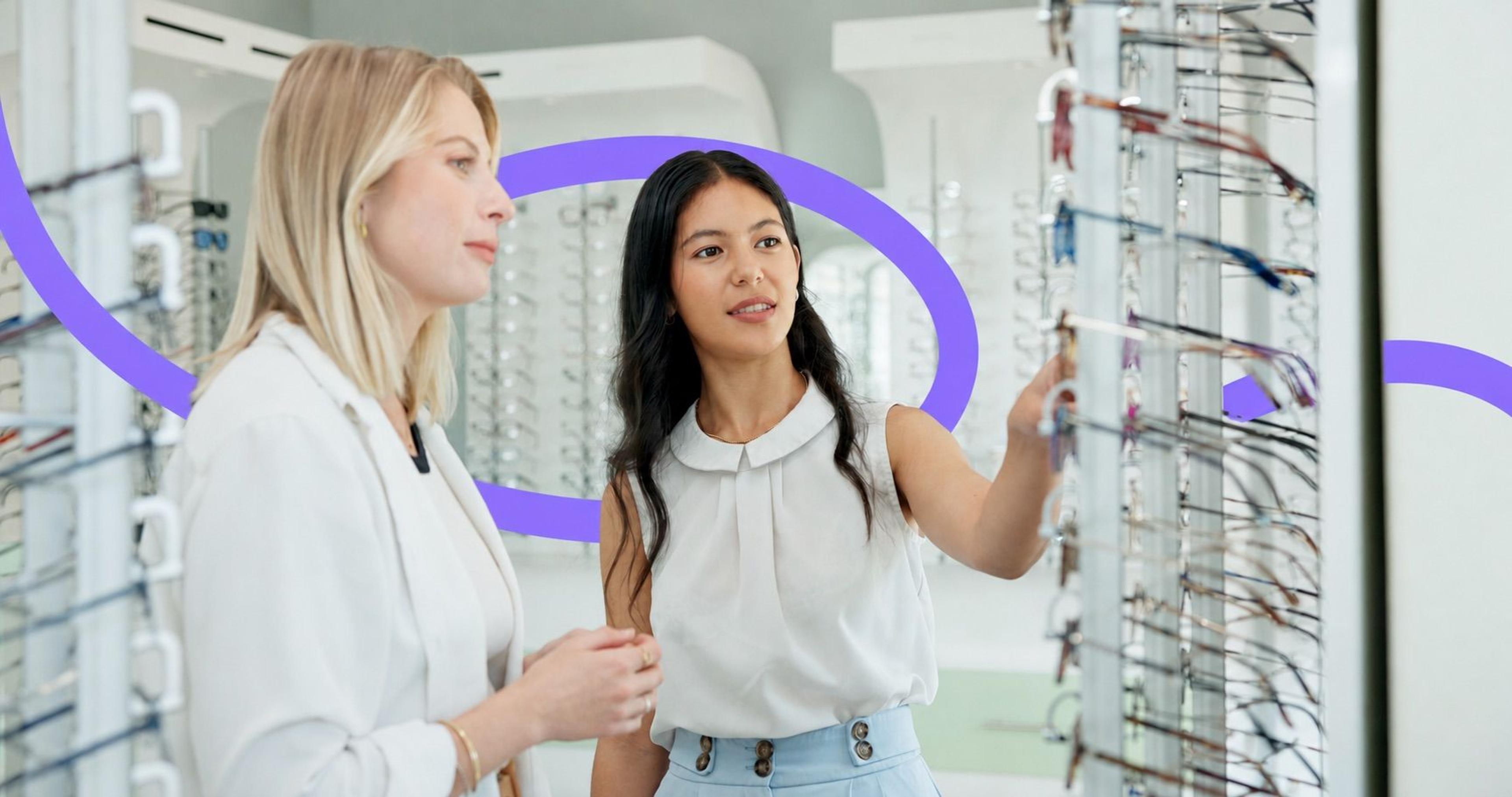 An optician helping a customer choose eyeglasses at a wall of frames