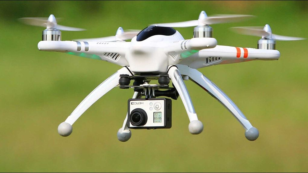A white quadcopter drone with four propellers is flying outdoors, equipped with a mounted GoPro camera underneath. The background is a blurred green field, indicating the drone is in motion.