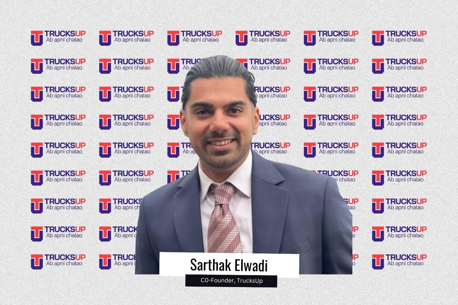 Sarthak Elwadi, Co-Founder of TrucksUp, is seen wearing a formal suit and smiling against a branded TrucksUp backdrop featuring the company’s logo and tagline “Ab apni chalao” in a repeated pattern.