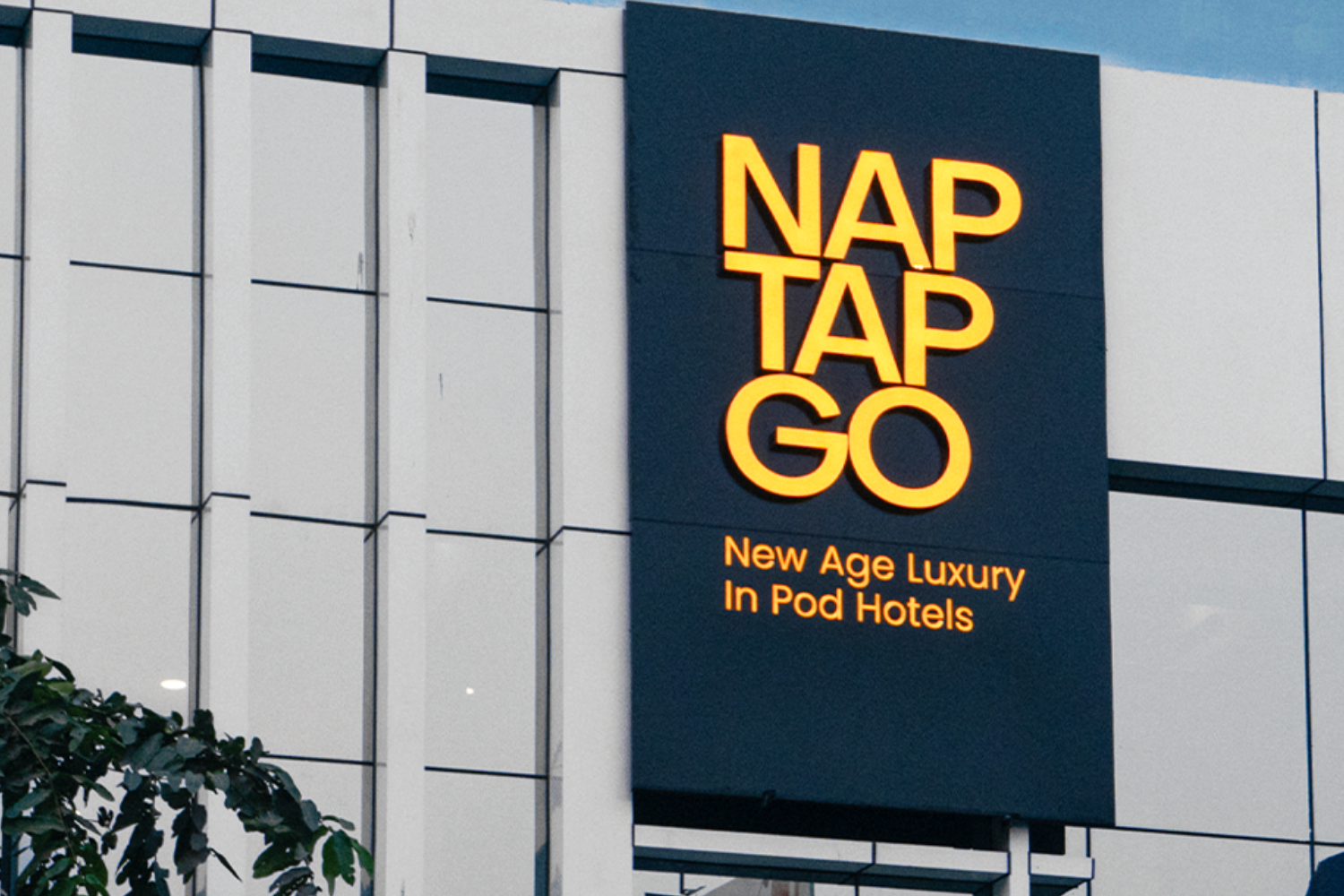 A sleek building facade with a glowing yellow signboard reading "NAP TAP GO" and the tagline "New Age Luxury in Pod Hotels," signifying modern accommodations.
