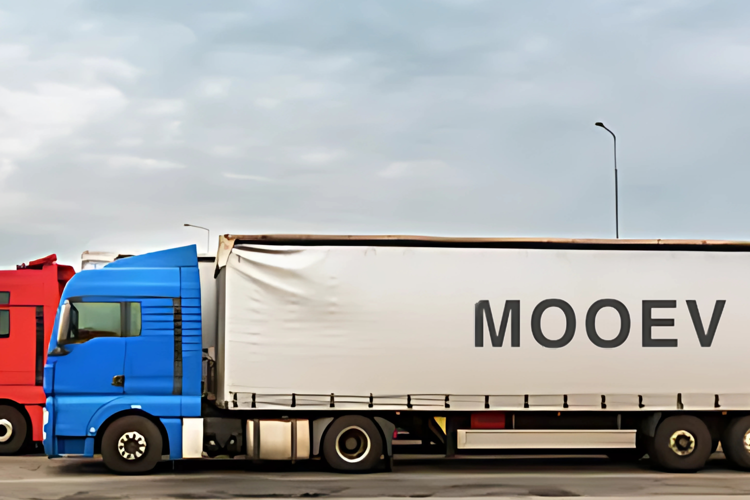 A large blue and white cargo truck parked side by side with a red truck. The trailer of the blue truck has the word "MOOEV" printed in bold letters. The background is overcast with a cloudy sky.