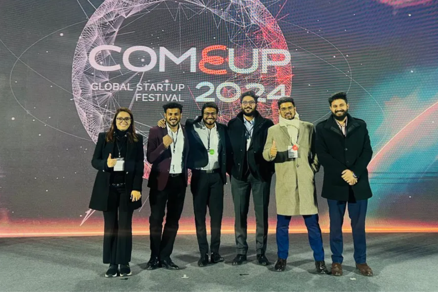 A group of six individuals dressed in formal and winter attire pose confidently on a stage at the "COMEUP Global Startup Festival 2024." A futuristic graphic with a glowing globe and network connections serves as the backdrop.