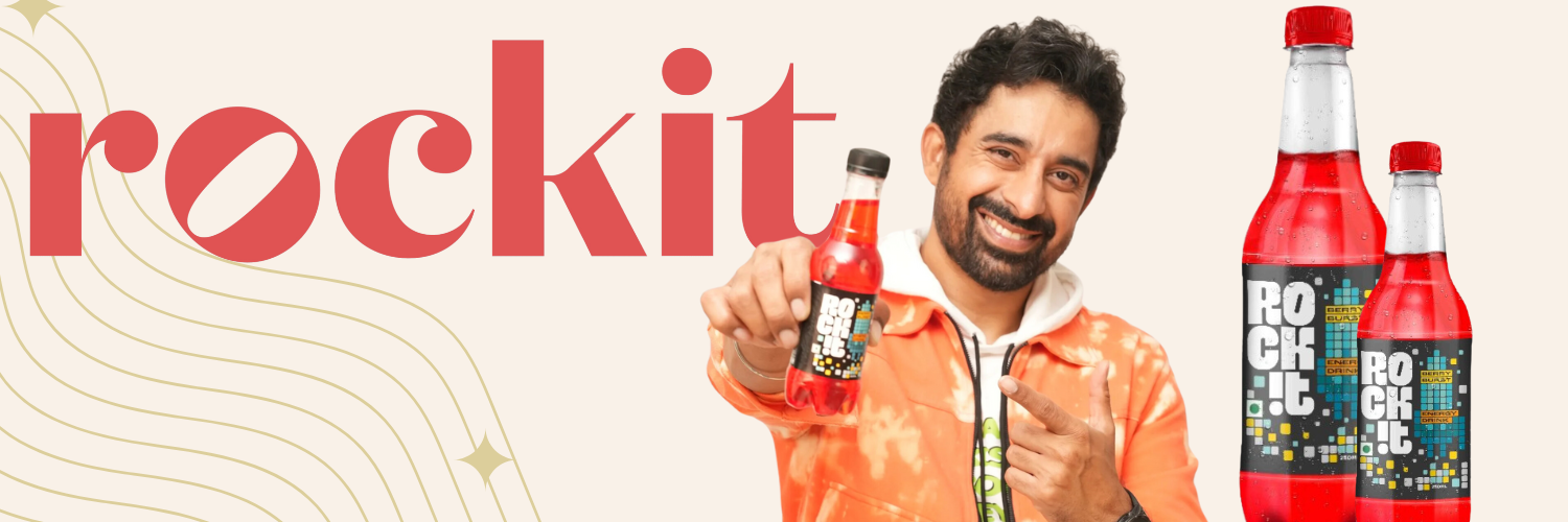 Investor Rannvijay Singha holding a bottle of Rockit, a berry burst energy drink, smiling and pointing at the bottle. The image features the Rockit logo prominently displayed on the left side, with additional bottles of Rockit on the right side of the image. The background is light with subtle golden wave patterns and star-like designs.