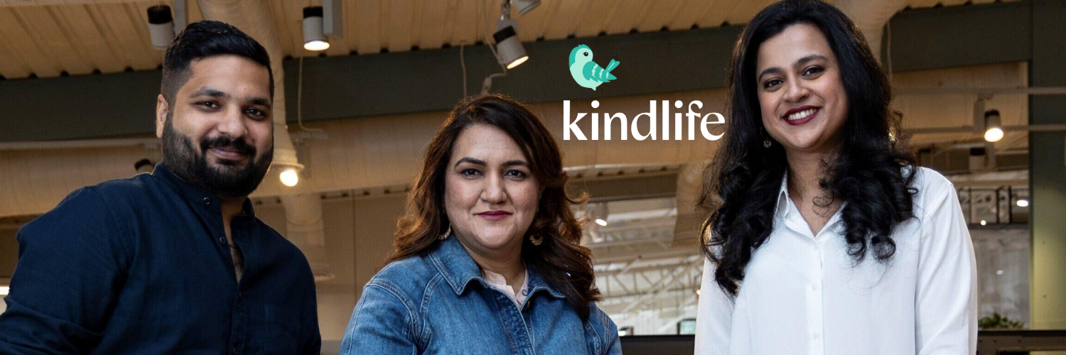  The image shows three individuals, likely the co-founders of Kindlife, standing together and smiling. The person on the left is wearing a dark blue shirt, the person in the center is wearing a denim jacket, and the person on the right is wearing a white blouse. Above them is the Kindlife logo, which features a small green bird icon. The setting appears to be a modern indoor space, possibly an office or a workspace, with lights and industrial design elements visible in the background.