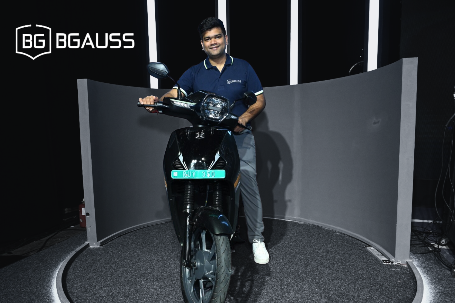 A man wearing a navy blue BGaus-branded polo shirt poses next to a sleek black electric scooter with the license plate 'RUV 350.' The setup is indoors, with a curved gray panel behind him and bright studio lighting illuminating the scene. The BGaus logo is displayed in the top-left corner