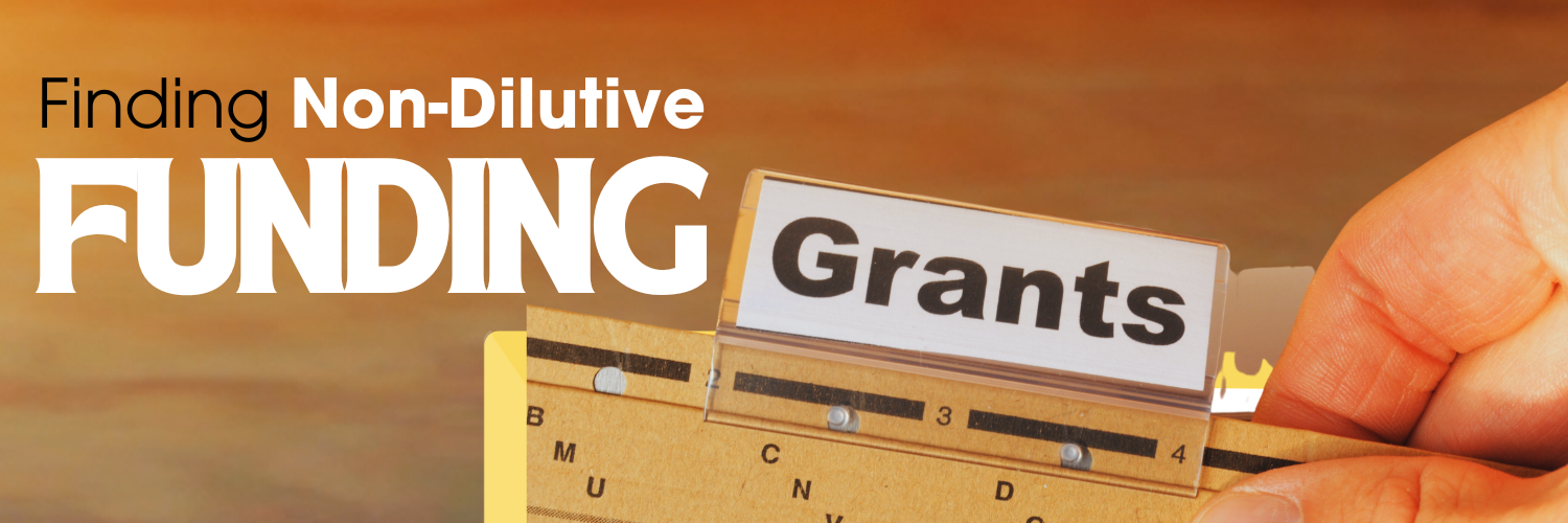 A banner titled "Finding Non-Dilutive Funding" featuring a close-up image of a hand pulling out a card labeled "Grants" from an organized file system. The background is warm and gradient, with the text "Finding Non-Dilutive Funding" prominently displayed in bold, white, and black letters on the left side.