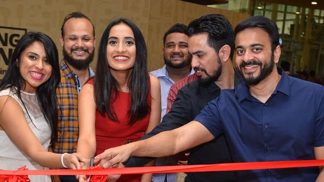 : Vineeta Singh at a store opening ceremony, dressed in a red knee-length dress. She is cutting a red ribbon with a large pair of scissors, surrounded by a group of five people who are cheering and smiling. The group includes both men and women, casually dressed, sharing the moment of celebration with her.