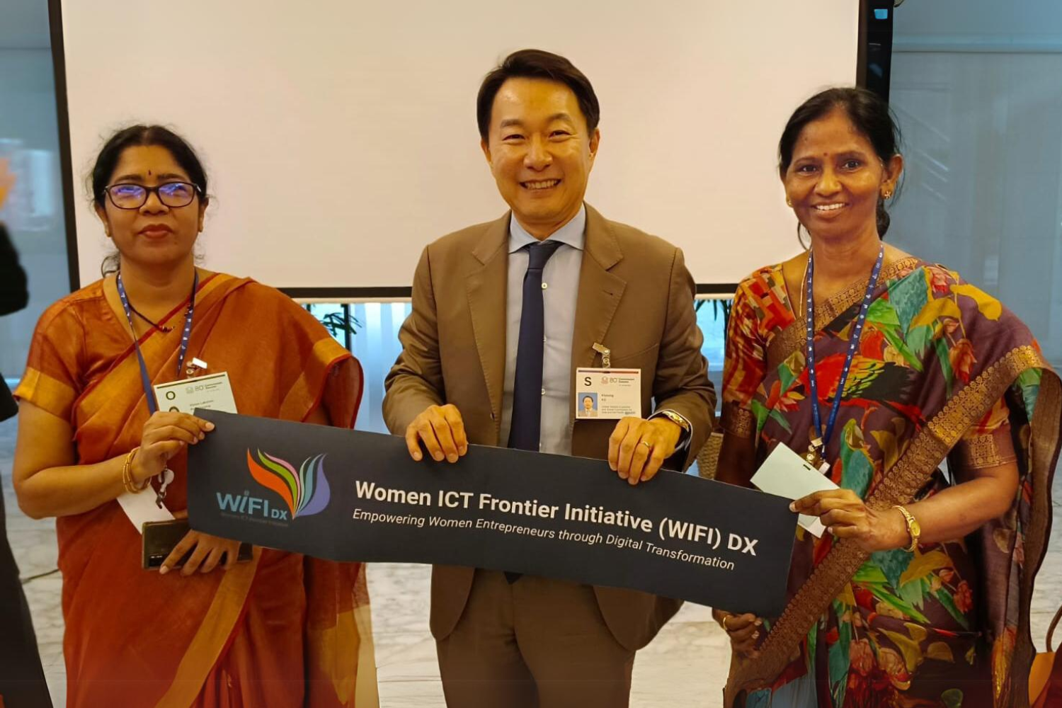 Three individuals holding a banner that reads 'Women ICT Frontier Initiative (WIFI) DX: Empowering Women Entrepreneurs through Digital Transformation.' The two women on each side are dressed in traditional sarees, while the man in the center is wearing a suit and smiling.