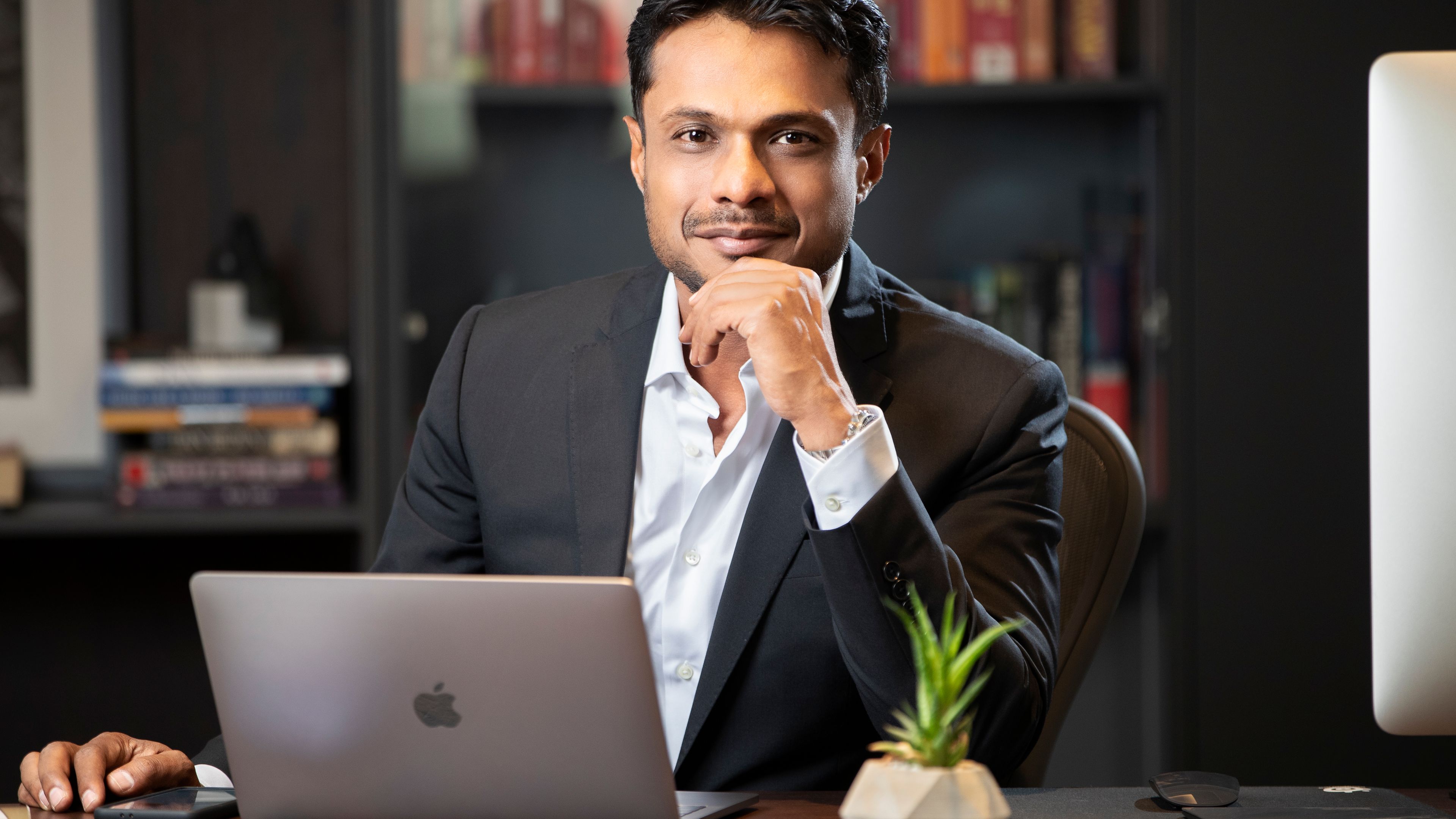 Sachin Bansal, dressed in a formal suit, is seated confidently at his desk with a laptop open in front of him, lightly resting his chin on his hand. Bookshelves in the background suggest a professional and intellectual setting.